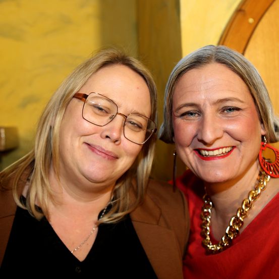 Zwei Frauen lächeln und posieren für ein Foto. Die Frau auf der linken Seite trägt eine Brille und eine Halskette. Die Frau auf der rechten Seite trägt eine Halskette und Ohrringe. Sie stehen eng beieinander.