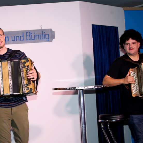 Zwei Männer spielen auf der Bühne, beide halten Akkordeons. Ein Mann steht vorne, trägt ein gestreiftes Hemd und Khaki-Hosen. Der andere Mann steht hinter einem Tisch, gekleidet in einem schwarzen T-Shirt und Jeans. Ein Banner mit der Aufschrift 'und Bildung' ist an der Wand hinter ihnen angebracht.
