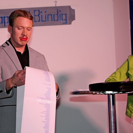 Ein Mann in einem grauen Anzug hält ein rosa Stück Papier und liest daraus. Die Wand hinter ihm hat ein blaues Schild mit weißem Text. Eine weitere Person in einer grünen Jacke ist teilweise sichtbar.