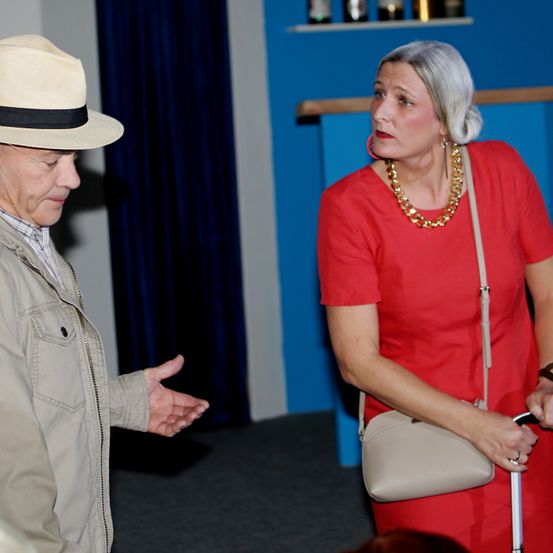 Ein älterer Mann mit Hut gestikuliert und spricht mit einer Frau in einem roten Kleid. Die Frau hält einen Stock und schaut zu. Sie stehen vor einem blauen Hintergrund.
