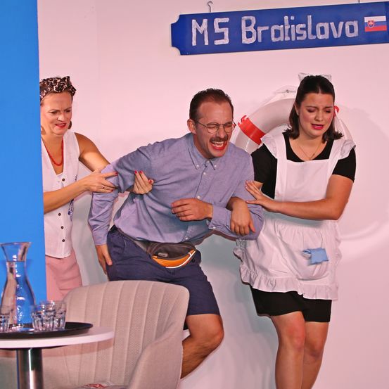 Drei Menschen lachen zusammen, einer hält eine Tasche, zwei Frauen in weißen Schürzen und ein Mann in einem blauen Hemd und Shorts. Sie stehen vor einer rosa Wand mit einem blauen Schild, auf dem 'MS Bratislava' steht. Ein Stuhl und ein Tisch mit Gläsern und einer Karaffe sind im Vordergrund.