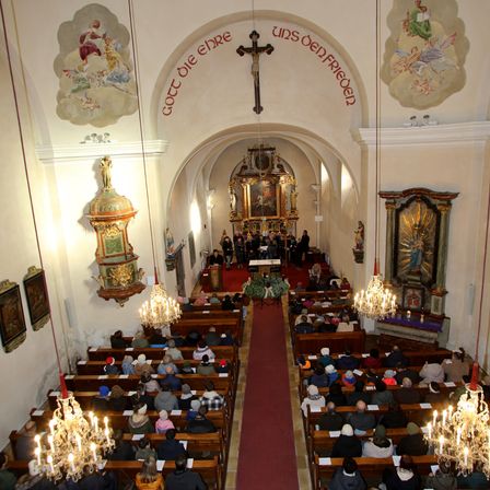 Ein Kircheninneres mit Menschen, die auf hölzernen Bänken sitzen, einem Chor, der vor einem Altar singt, einem roten Teppich, der zum Altar führt, und dekorativen Kronleuchtern und religiösen Kunstwerken.