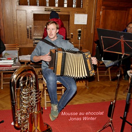Ein Mann spielt Akkordeon, während er auf einem Stuhl sitzt. Vor ihm steht ein Tuba, daneben ein Notenständer. Hinter ihm steht eine Person in einem roten Mantel.