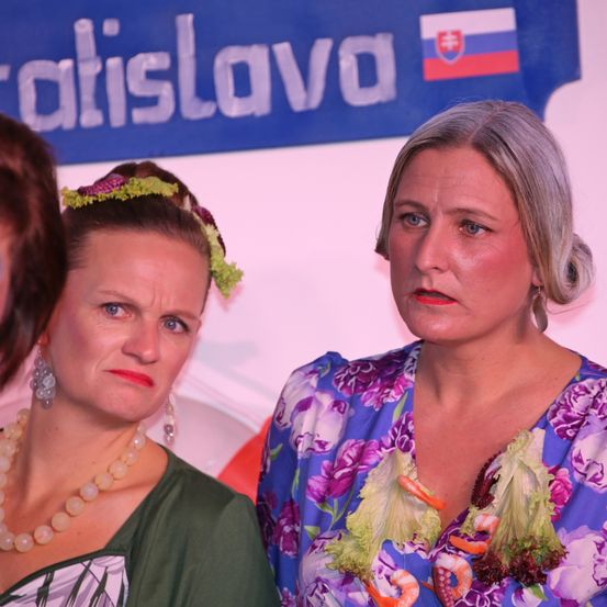 Zwei Frauen stehen nebeneinander, eine trägt ein Blumenkleid, das mit Salatblättern und Garnelen verziert ist, die andere ein grünes Kleid mit einer Blumenkrone. Sie schauen beide direkt in die Kamera. Im Hintergrund ein Banner mit dem Wort Bratislava und der slowakischen Flagge.