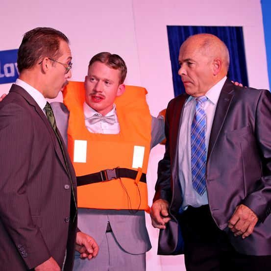 Drei Männer stehen in formeller Kleidung; einer trägt ein orangefarbenes Rettungsweste und eine Fliege. Der Mann links trägt eine Brille, der Mann rechts trägt eine karierte Krawatte. Sie scheinen sich in einem professionellen Umfeld zu befinden.