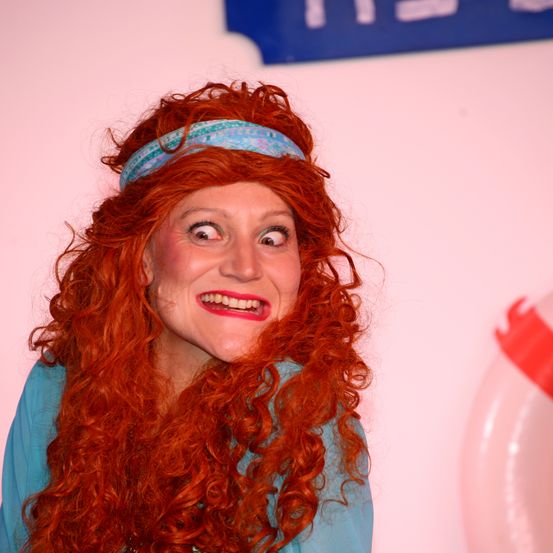 Eine Frau mit roten Locken und einem blauen Stirnband lächelt und scheint für ein Foto zu posieren. Sie trägt ein blaues Oberteil.