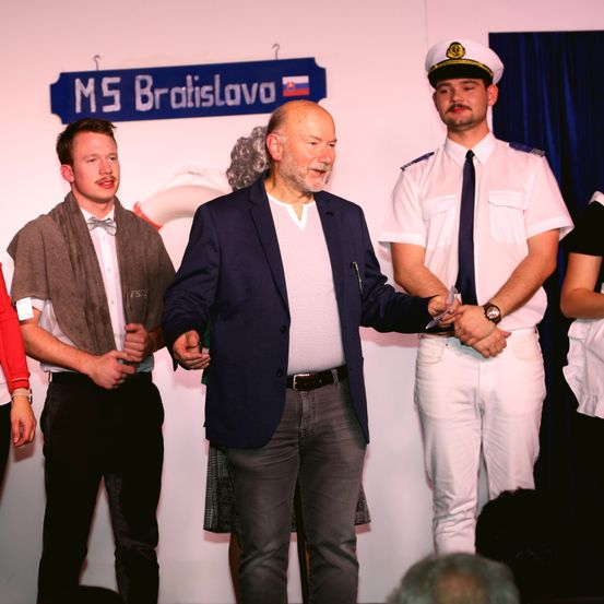 Drei Männer stehen auf einer Bühne, einer hält eine Schere, möglicherweise für eine Schneidezeremonie. Der Mann in der Mitte trägt einen marineblauen Blazer und graue Hosen. Ein blaues Schild mit 'MS Bratislava' ist an der Wand hinter ihnen angebracht.