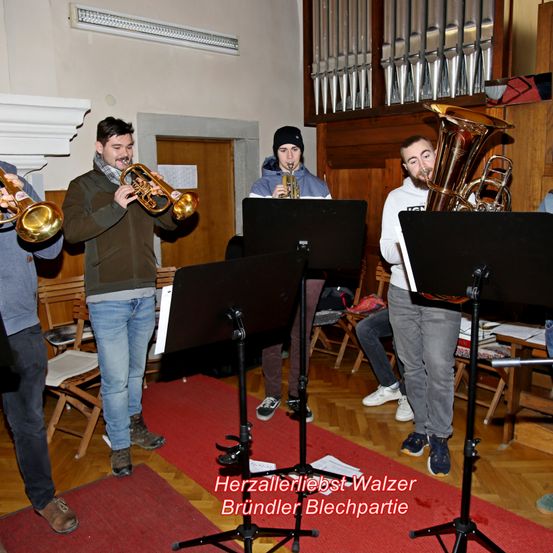 Eine Gruppe von Musikern in einem Raum mit Holzböden, einer spielt Trompete, ein anderer Posaune, und andere beobachten. Es gibt Notenständer und Stühle um sie herum. Der Text lautet Herzallerliebst Walzer Brundler Blechparteie.