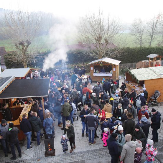 Eine Menschenmenge, möglicherweise auf einem Markt, versammelt sich um Stände und einen Feuerkorb. Einige tragen Mützen, Brillen und Schuhe. Es gibt Bäume und einen Zaun in der Gegend. Die Stände sind aus Holz und einige sind mit grünem Tuch bedeckt. In der Ferne gibt es Häuser und ein Feld.