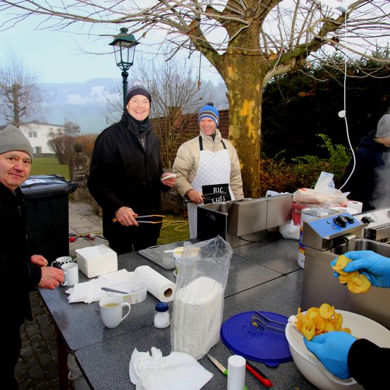 Vier Personen stehen um einen Tisch herum und bereiten Essen zu. Zwei Männer tragen Schürzen, eine mit 'BIG CHEF' darauf. Es gibt eine Schüssel mit Essen, Papierhandtücher und Tassen auf dem Tisch.