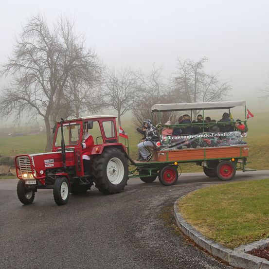 Ein roter Traktor zieht einen grünen Anhänger mit Passagieren auf einer nebligen Straße. Der Traktor hat eine Flagge an seiner Seite.