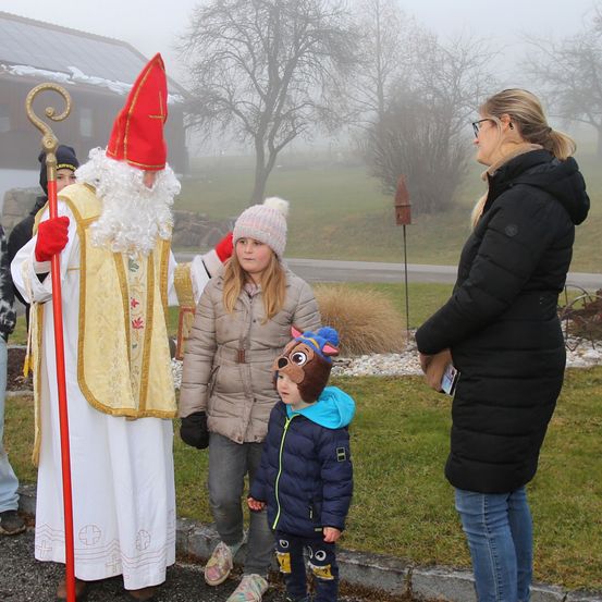 Eine Gruppe von Menschen steht an einem nebligen Tag im Freien. Ein Mann, der als Sankt Nikolaus gekleidet ist, hält einen Stab. Zwei Kinder stehen vor ihm, eines trägt einen Bärenhut. Eine Frau in einer schwarzen Jacke steht rechts.