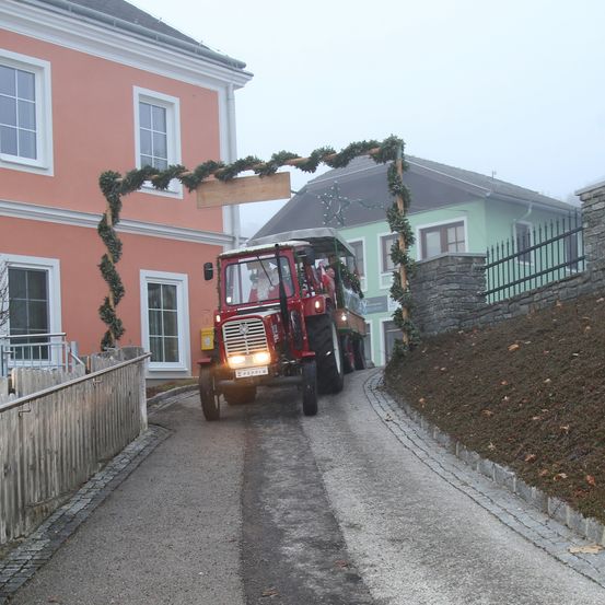 Ein roter Traktor fährt auf einer nebligen Straße vorbei und durchfährt einen mit Grün bekränzten Torbogen. Ein Haus mit rosa Wänden und ein grün gestrichenes Haus befinden sich auf beiden Seiten.