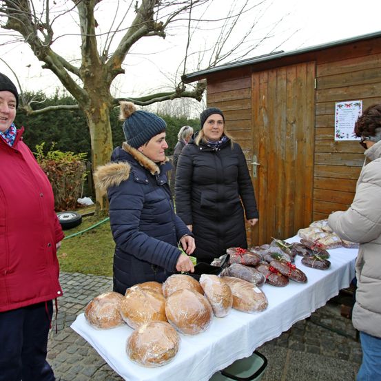 Vier Frauen stehen vor einem Holzschuppen. Eine Frau wickelt Brot auf einem Tisch ein. Andere Frauen schauen zu. Bäume und ein Schild sind im Hintergrund.