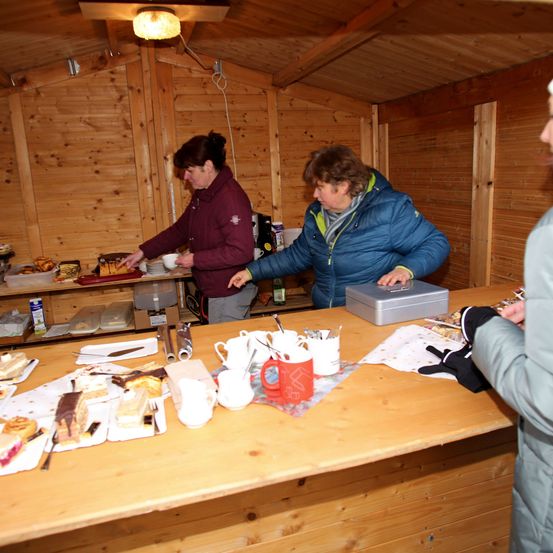 Zwei Frauen in einer kleinen Holzhütte bereiten Essen zu. Der Tisch ist mit Desserts, Tassen und Tellern gefüllt. Eine Lampe ist an der Decke montiert.