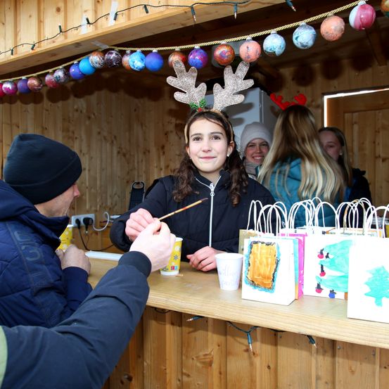 Ein Mädchen mit Hirschgeweih-Kopfband lächelt in einer winterlichen Umgebung, hält einen Pinsel. Hinter ihr sehen Leute zu, und Papiertüten mit Kunstwerken stehen auf dem Tresen. Ein Mann sitzt in der Nähe.