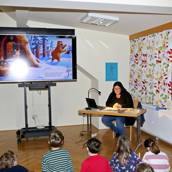 Eine Frau liest an einem Schreibtisch vor einem Bildschirm mit einer Kindergeschichte. Kinder sitzen auf dem Boden und schauen zu.