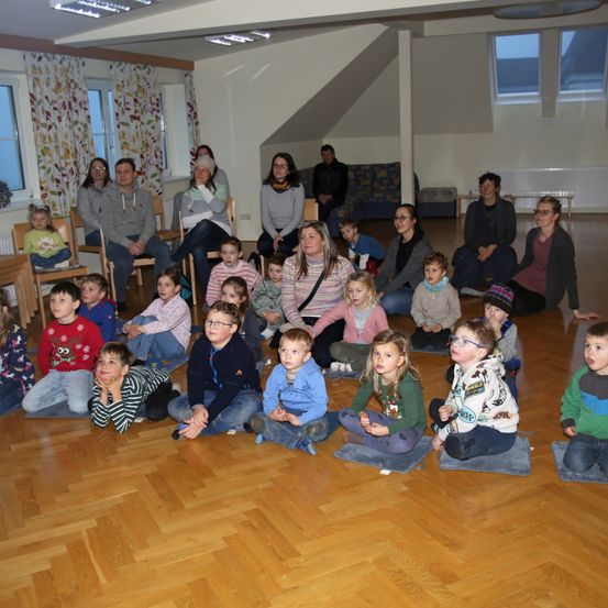 Eine Gruppe von Kindern und Erwachsenen sitzt auf einem Holzboden, wahrscheinlich in einem Klassenzimmer. Einige tragen Brillen und andere Mützen. Es gibt Fenster mit Vorhängen und Lampen an der Decke.
