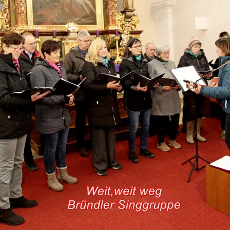 Ein Chor von Erwachsenen in Winterkleidung singt in einer Kirche. Eine Frau vorne spielt Gitarre. Der Boden ist mit rotem Teppich bedeckt.