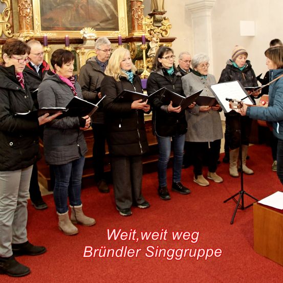 Ein Chor von Erwachsenen in Winterkleidung singt in einer Kirche. Eine Frau vorne spielt Gitarre. Der Boden ist mit rotem Teppich bedeckt.