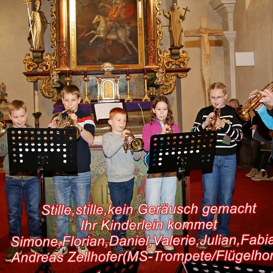 Sechs Kinder in einer Kirche spielen Trompeten. Der Altar hinter ihnen ist mit Statuen und einem Kreuz geschmückt. Die Kinder tragen Freizeitkleidung. Der rote Teppich hat den Text 'Stille, stille, kein Gerausch gemacht Ihr Kinderlein kommt'.