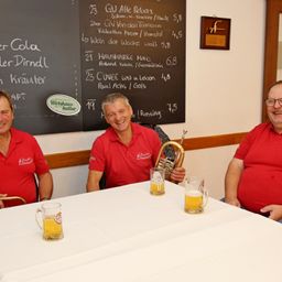 Drei Männer sitzen an einem Tisch mit Gläsern Bier. Hinter ihnen steht eine Tafel mit einer Speisekarte in deutscher Sprache.
