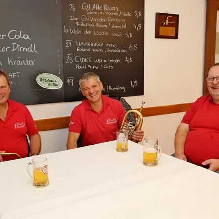 Drei Männer sitzen an einem Tisch mit Gläsern Bier. Hinter ihnen steht eine Tafel mit einer Speisekarte in deutscher Sprache.