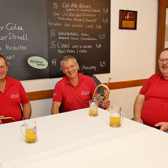 Drei Männer sitzen an einem Tisch mit Gläsern Bier. Hinter ihnen steht eine Tafel mit einer Speisekarte in deutscher Sprache.