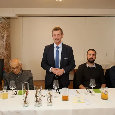 Vier Männer sitzen um einen Tisch herum, drei vorne und einer dahinter. Der Mann im Anzug trägt einen Sakko und eine Krawatte. Der Tisch hat Gläser, Servietten und Bier. Dahinter befinden sich weiße Türen.