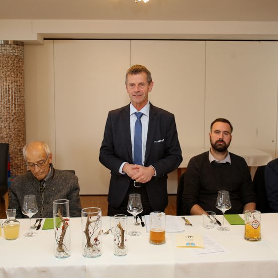 Vier Männer sitzen um einen Tisch herum, drei vorne und einer dahinter. Der Mann im Anzug trägt einen Sakko und eine Krawatte. Der Tisch hat Gläser, Servietten und Bier. Dahinter befinden sich weiße Türen.