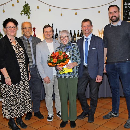 Eine Gruppe von Menschen steht in einem Raum mit Weinflaschen im Hintergrund. Eine Frau hält einen Blumenstrauß und ein Buch.