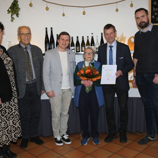 Eine Gruppe von Menschen steht in einem Raum zusammen, eine Frau hält einen Blumenstrauß und ein Zertifikat. Hinter ihnen steht ein Tisch mit Flaschen auf einem Regal.