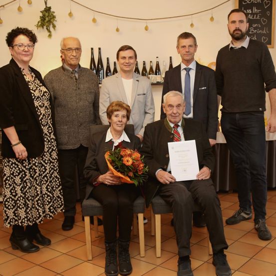 Eine Gruppe von Menschen steht und sitzt in einem Raum. Eine Frau in der Mitte hält Blumen und lächelt. Ein Mann vorne hält ein Zertifikat. Flaschen sind dahinter angeordnet. Eine Tafel ist an der Wand montiert.
