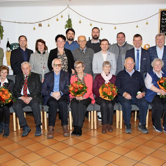 Eine Gruppe von Menschen sitzt auf Stühlen in einem Raum, einige halten Blumen. Sie sind alle in formeller Kleidung gekleidet. Hinter ihnen ist eine Tafel an der Wand montiert und einige Pflanzen hängen an der Decke. Es gibt Flaschen auf einem Regal.
