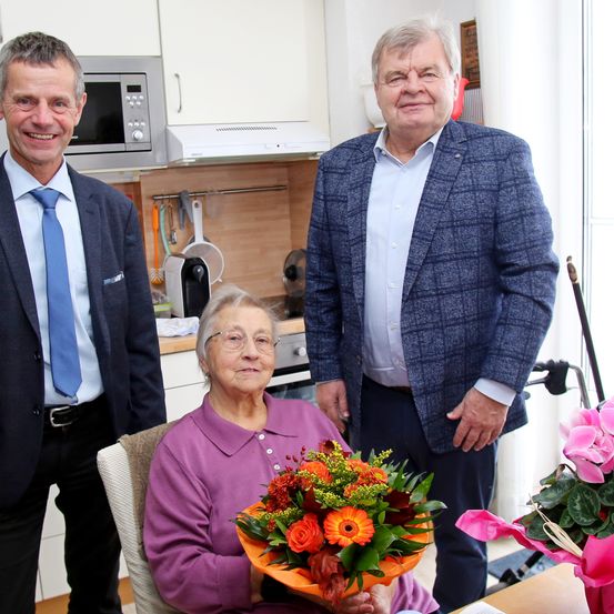 Drei Personen stehen in einer Küche. Eine Frau auf einem Stuhl hält einen Blumenstrauß. Ein Mann hinter ihr lächelt und ein anderer Mann auf der linken Seite schaut in die Kamera.