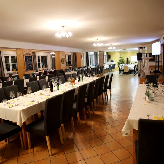 Ein großer Speisesaal ist für eine formelle Veranstaltung vorbereitet. Lange Tische sind mit weißen Tischdecken bedeckt, und Weinflaschen und Gläser sind arrangiert. Stühle sind ordentlich aufgereiht. Ein Bildschirm und Lautsprecher sind im Hintergrund aufgestellt.