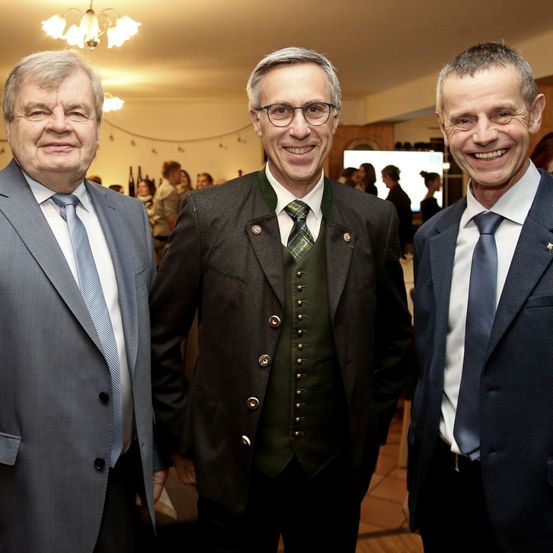 Drei Männer in Anzügen stehen eng beieinander und lächeln, während sie für ein Foto posieren. Hinter ihnen stehen einige Leute, und eine Kronleuchter hängt von der Decke.