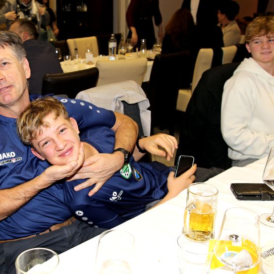 Ein Mann und ein Junge sitzen an einem Tisch und halten Bier. Der Mann hat seinen Arm um den Jungen. Ein anderer Junge steht hinter ihnen. Sie sind in einem Restaurant mit anderen Leuten.