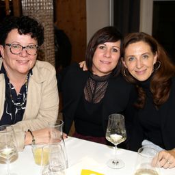 Drei Frauen sitzen zusammen an einem Tisch und lächeln für ein Foto. Die Frau links trägt eine Brille und eine Halskette. Die Frau in der Mitte trägt ein schwarzes Oberteil mit Muster. Die Frau rechts trägt einen Ring und Ohrringe. Gläser, Tassen und Weingläser stehen auf dem Tisch.