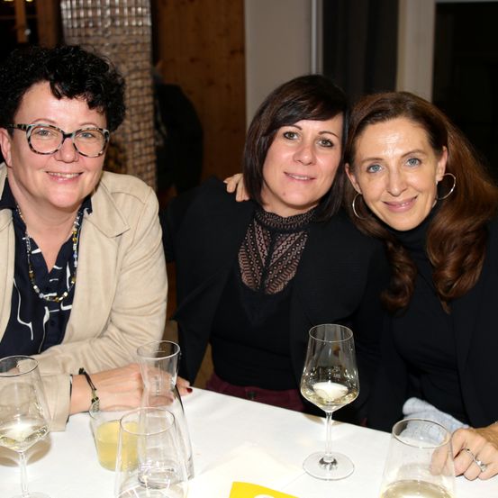 Drei Frauen sitzen zusammen an einem Tisch und lächeln für ein Foto. Die Frau links trägt eine Brille und eine Halskette. Die Frau in der Mitte trägt ein schwarzes Oberteil mit Muster. Die Frau rechts trägt einen Ring und Ohrringe. Gläser, Tassen und Weingläser stehen auf dem Tisch.