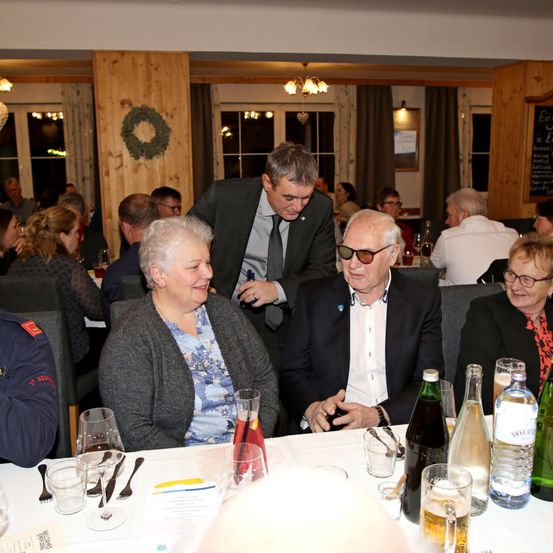 Eine Innenveranstaltung mit mehreren Personen, die an einem Tisch sitzen. Ein Mann in Anzug und Krawatte steht und spricht mit zwei Männern in Anzügen. Auf dem Tisch stehen Gläser, Flaschen und Besteck. Der Raum hat Holzwände und Lampen.