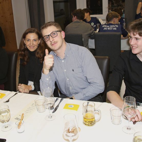 Ein Mann gibt einen Daumen hoch, während er mit einer Frau und einem anderen Mann an einem Tisch sitzt. Hinter ihnen sitzen zwei Personen an einem anderen Tisch. Leere Gläser, Teller und Besteck sind auf dem Tisch.