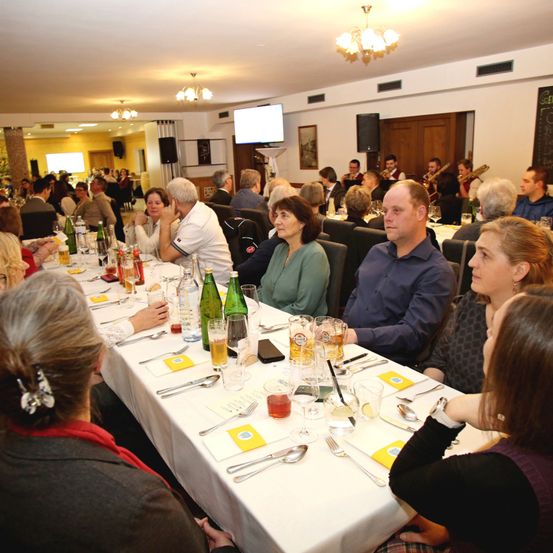 Eine Gruppe von Menschen sitzt an einem langen Tisch in einem Essbereich, mit Flaschen, Gläsern und Besteck. Eine Band spielt im Hintergrund.