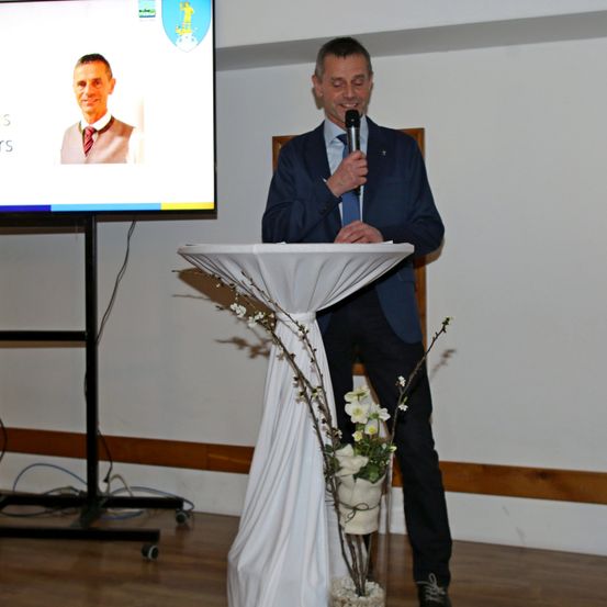 Ein Mann spricht an einem Podium in einem Raum mit einem großen Bildschirm, der ein Bild eines Mannes zeigt.