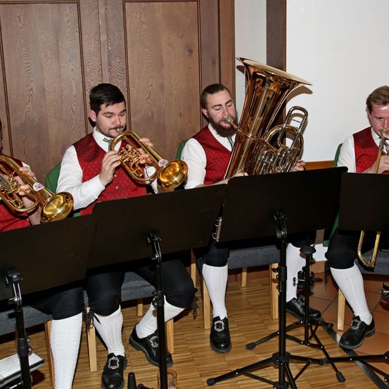 Vier Männer in roten Westen und weißen Socken sitzen und spielen Blechblasinstrumente in einem Raum. Einer spielt Trompete, ein anderer Tuba und die anderen zwei Posaunen. Sie werden von Notenständern begleitet.