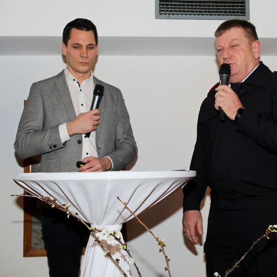 Zwei Männer stehen vor einem Podium, jeder hält ein Mikrofon in der Hand. Der Mann auf der linken Seite trägt einen grauen Anzug und scheint zu sprechen. Der Mann auf der rechten Seite ist in einem schwarzen Uniform.