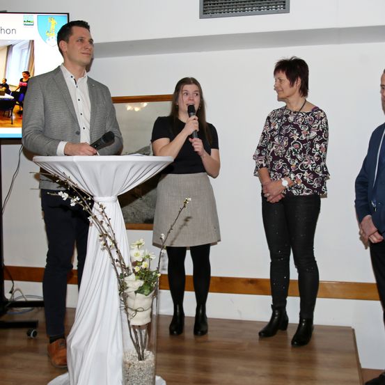 Eine Gruppe von Menschen steht auf einer Bühne mit einem Mikrofon und Blumen. Eine Frau spricht, während eine andere zuhört. Ein Mann steht hinter einem Podium. Ein Bildschirm zeigt ein Bild an.