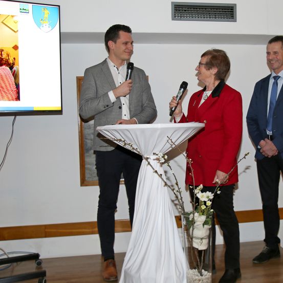 Ein Mann und eine Frau halten Mikrofone, während sie vor einem Blumenpodium stehen. Dahinter lächelt ein Mann. Eine Leinwand mit einem Bild ist an der Wand montiert.