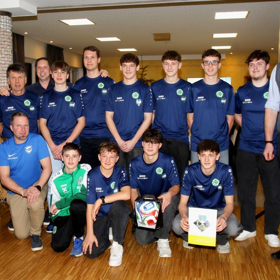 Eine Gruppe junger Männer und ältere Erwachsene posiert für ein Foto. Sie tragen blaue Uniformen mit grünen Logos und einige halten einen Pokal. Sie stehen auf einem Holzboden.