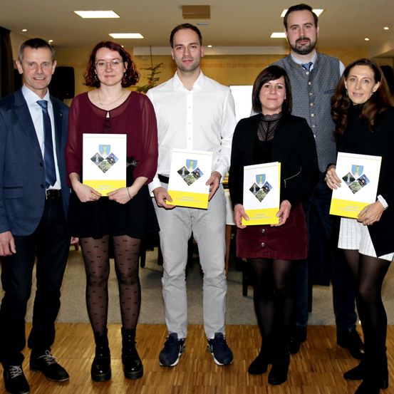 Eine Gruppe von sechs Personen steht zusammen in einem Raum, lächelt und posiert für ein Foto. Sie alle halten gelbe Broschüren mit demselben Design.